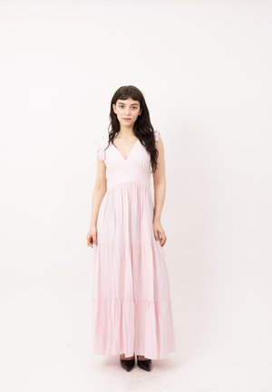 Mujer con cabello oscuro y largo que lleva un vestido maxi sin mangas de color rosa claro con volantes y zapatos negros puntiagudos, de pie frente a un fondo blanco liso.