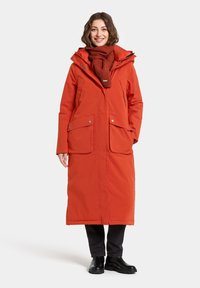 Long, bright orange coat with a hood, featuring two front pockets, snap buttons, and a textured red scarf. Worn with black pants and boots.
