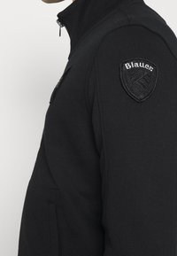 Svart zip-up sweatshirt med en strukturerad tyg, ståkrage och en framträdande svart broderad lapp på vänster axel.