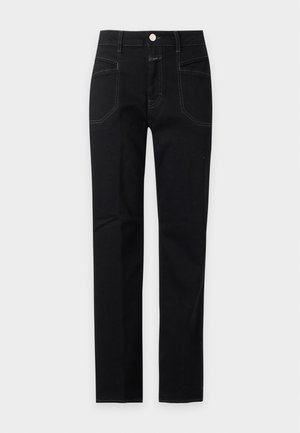 Femme debout portant un polo gris à manches longues, un jean taille haute noir et des mocassins marron foncé sur un fond blanc uni.