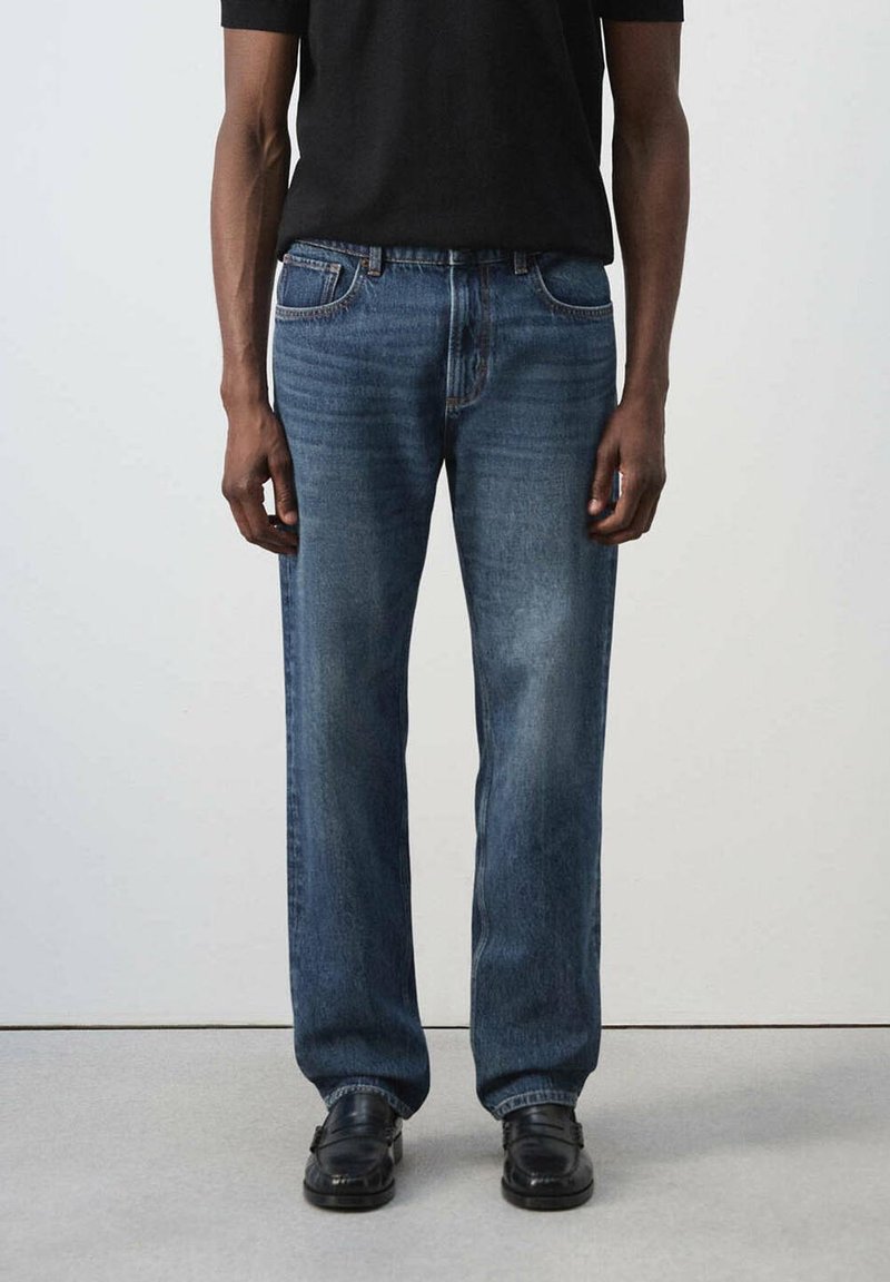 Personne portant un jean bleu droit, des chaussures noires et une chemise noire à manches courtes, debout devant un fond uni.