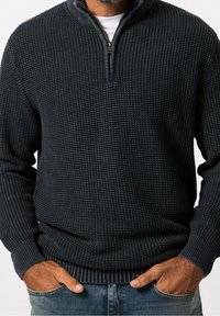Sweater texturé marine avec une demi-fermeture éclair, des poignets et un ourlet côtelés, arborant un motif tissé. Porté par-dessus un t-shirt blanc.