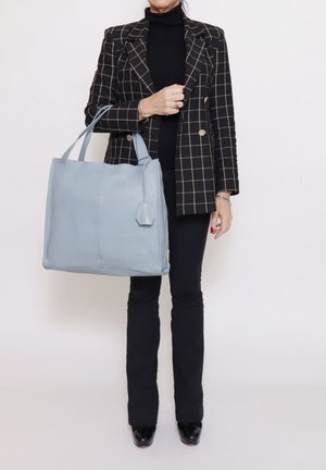 Sac fourre-tout en cuir bleu clair avec une forme structurée, des poignées supérieures et un détail d'étiquette sur le côté, associé à un blazer à carreaux noir et blanc.