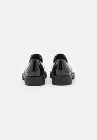 Paire de chaussures de ville en cuir noir brillant vues de dos sur un fond blanc avec des semelles épaisses noires.