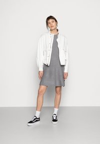 Balta bomber jakna virs pelēka pieguļoša kleita, melnbaltas kedas, baltas zeķes, stāv uz gaiši pelēka grīdas pret vienkāršu fona.