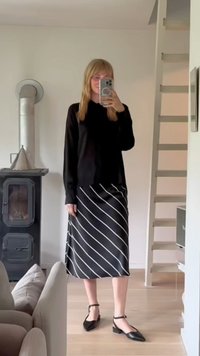 Černý dlouhý rukáv top spárován s černou sukní s bílými diagonálními pruhy. Modelka nosí černé ploché boty se zápěstními pásky.