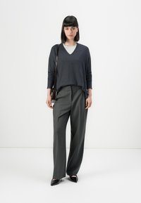 Donna con capelli neri, che indossa un maglione grigio scuro con scollatura a V, una maglietta grigia chiara sotto, pantaloni larghi grigi e scarpe nere a punta, mentre porta una borsa a tracolla scura.