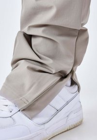Pantalones beiges ajustados con puños con cremallera, combinados con unas zapatillas blancas. La tela tiene una textura suave con sutiles estrías verticales.