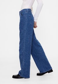 Sinisistä, leveälahkeisista farkuista, jotka on tehty teksturoidusta kankaasta ja joissa on hienovarainen kuvio. Niissä on perinteiset taskut ja napillinen suljenta, yhdistetty mustiin kenkiin.