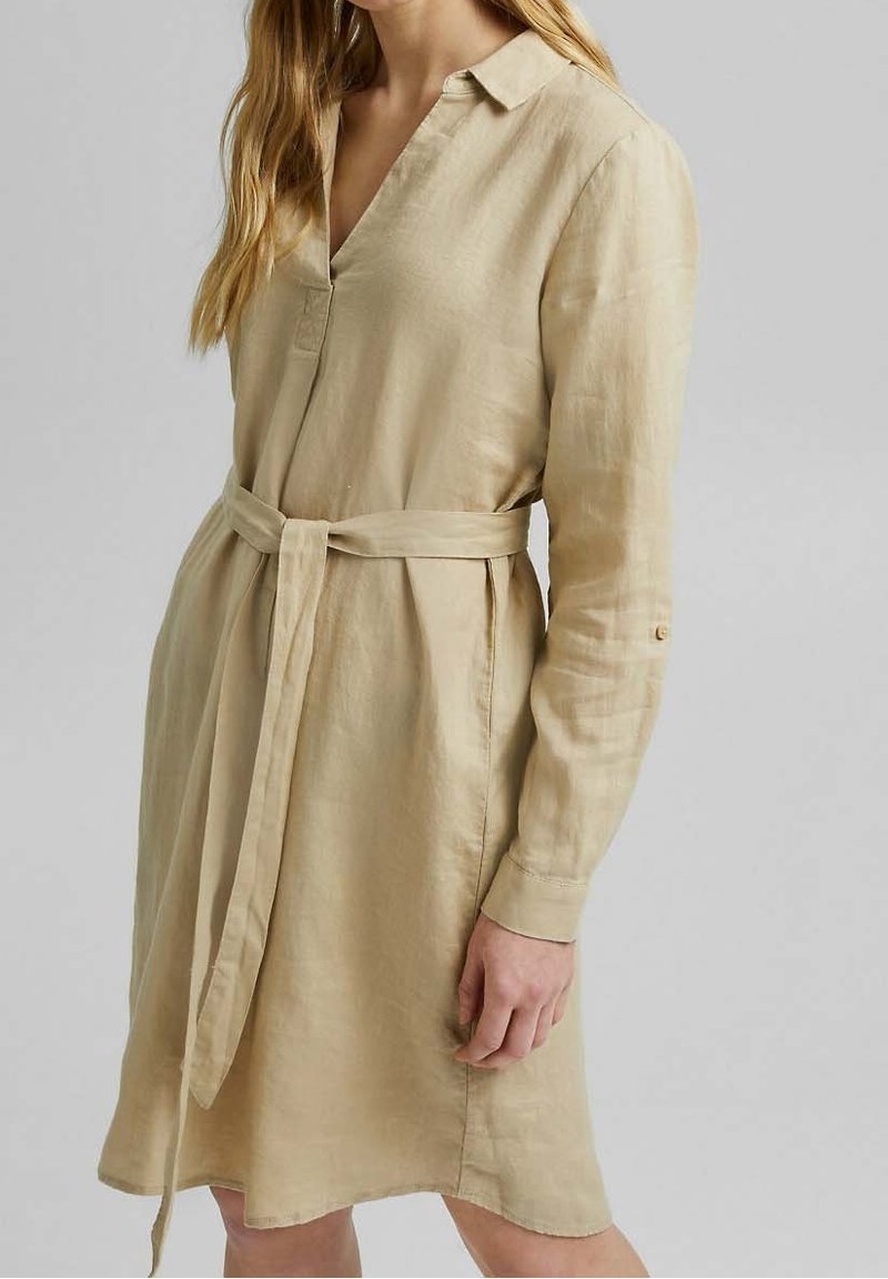 Femme portant une robe beige à manches longues avec col, décolleté en V et ceinture nouée à la taille, debout devant un fond uni.