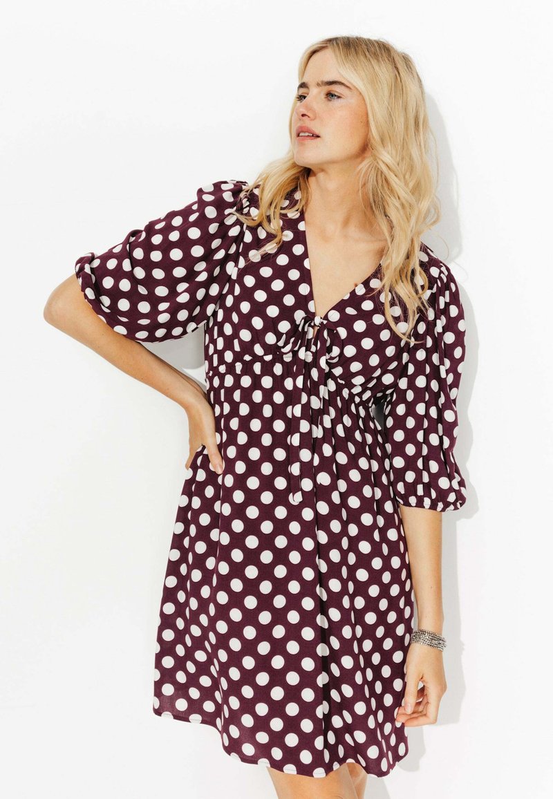 Robe bordeaux à pois avec des manches courtes bouffantes, un décolleté en V et une taille froncée. Présente un détail noué à l'avant et des poches latérales.