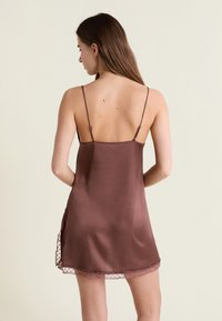 Mujer con cabello largo y castaño, vistiendo un vestido de satén marrón con tirantes delgados, vista desde atrás sobre un fondo liso y claro.