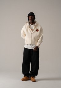 Κρεμ ζιπ-από hoodie με σχέδιο μήλου, λευκό μπλουζάκι, σκούρα φαρδιά τζιν παντελόνια και καφέ μποτάκια. Συμπληρώνεται με έναν μαύρο καπέλο και κολιέ.