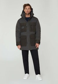 Mörkgrå vadderad parkasjacka med flera fickor, en dragkedja fram och avtagbar huva. Har svarta detaljer och elastiska ärmslut.