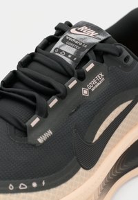 Baskets de course noires avec une tige en tissu texturé, marquage Gore-Tex et accents rose clair sur la semelle et les œillets. Présente un logo de swoosh Nike.