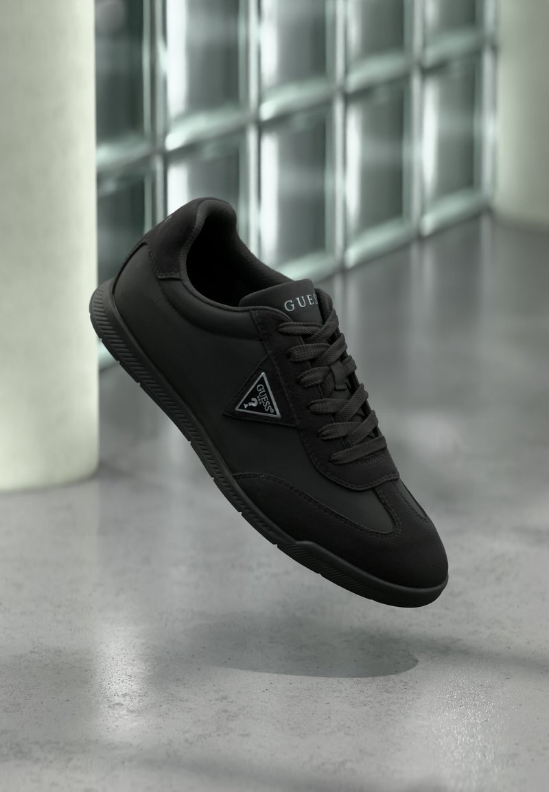 Baskets Guess noires avec lacets et logo sur le côté et la languette, flottant au-dessus d'un sol en béton poli près d'un mur en blocs de verre.