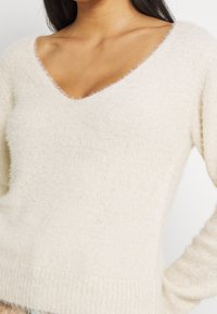 Hellbeige, flauschiger Pullover mit tiefem V-Ausschnitt, langen Ärmeln und geripptem Saum. Die Textur wirkt weich und hat ein leicht fusseliges Finish.