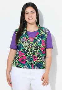 Mehrfarbige Weste mit einem auffälligen Tierdruckdesign, das grüne, pinke und schwarze Muster zeigt, über einem lila T-Shirt getragen, kombiniert mit weißen Hosen.