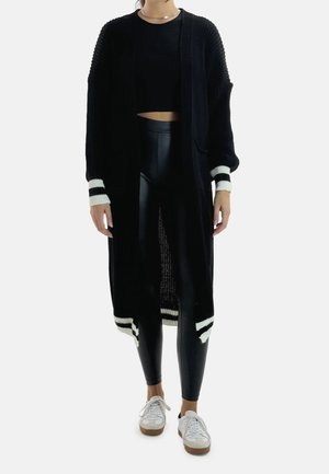 Kvinde iført sort lang cardigan med hvide striber på ærmerne og kanten, sort cropped top, sorte leggings og hvide sneakers.