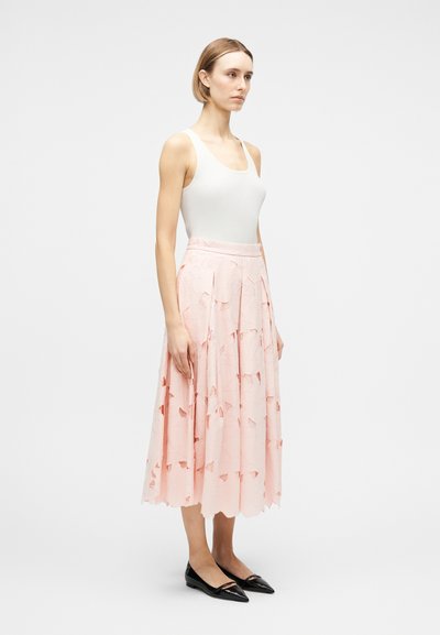 Γυναίκα με λευκό αμάνικο τοπ, ανοιχτό ροζ υφασμένο midi φούστα και μαύρα μυτερά φλατ παπούτσια, στέκεται πλάγια μπροστά σε απλό φόντο.