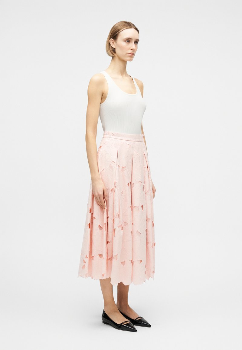 Donna che indossa una canottiera bianca, una gonna midi rosa pallido strutturata e ballerine nere con punta, in piedi di profilo su uno sfondo semplice.