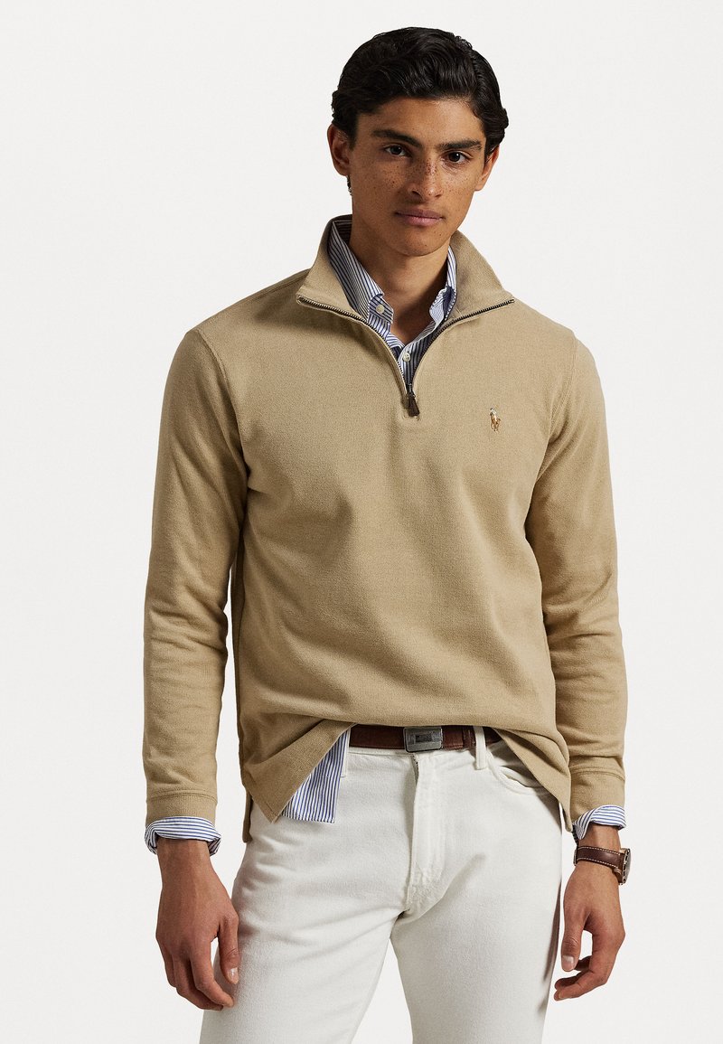 Beige zip-up sweater with ribbed texture, layered over a blue-and-white striped shirt, paired with white pants and a brown belt.
