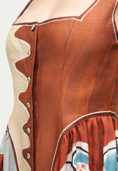 Κοντινή λήψη του κορσέ ενός φορέματος με σκουρόχρωμο και μπεζ ύφασμα, διακοσμητικά κουμπιά, κυματιστές άκρες και μέρος της φούστας με πιετά και πολύχρωμα μοτίβα.