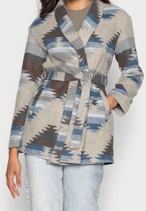 Kvinde iført en jakke med bælte med et blåt, gråt og brunt geometrisk mønster over lyse denimjeans.