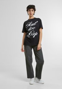 Camiseta de algodón negra con texto blanco que dice "Alma demasiado profunda", combinada con jeans gris oscuro y zapatillas blancas. Manga corta, corte relajado.