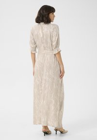 Robe maxi en beige clair avec un motif tourbillonnant, manches trois quarts, taille ceinturée et tissu fluide. Portée avec des chaussures à talons légers.