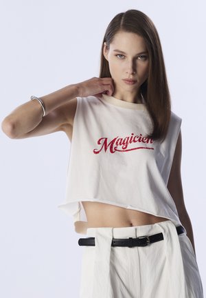 Camiseta blanca recortada con la palabra "Magicien" en rojo cursiva. El modelo la combina con pantalones blancos y un cinturón negro, presentando un diseño minimalista y suave.