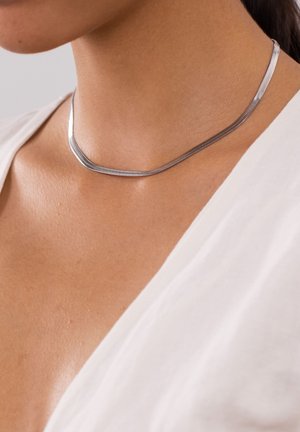 Collier chaîne serpent plate en argent porté autour du cou sur une peau claire, associé à un haut blanc à col en V.