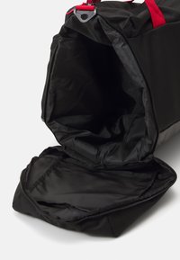 Schwarze Duffel-Tasche aus strapazierfähigem Stoff, mit roten Trägern. Das Innenleben ist geräumig, mit einer glatten, gefütterten Textur und einem Reißverschluss.