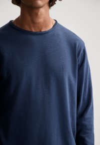 Tengeri kékszínű, hosszú ujjú póló, puha anyagból készült, kerek nyakkivágással és finom logódísszel a bal oldalon. Simás textúra, laza szabás.