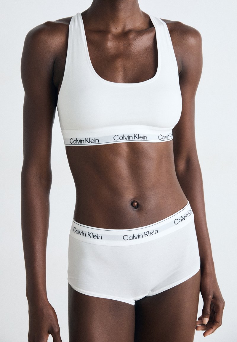 Bijeli set sportskog grudnjaka i gaćica Calvin Klein. Grudnjak ima izrez s niskim vratom, dok gaćice imaju pojas s logotipom. Glatka, rastezljiva pamučna tkanina.