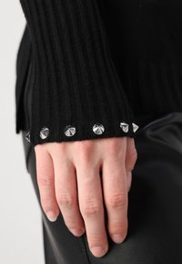 Pull en maille côtelée noir avec des clous en pyramide argentés le long du poignet. La main reposant sur une surface noire contraste avec le tissu.