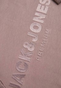 Tela de color malva claro con texto bordado texturizado que dice "JACK&JONES PREMIUM" en un tono contrastante. Textura suave y lisa, estilo casual.