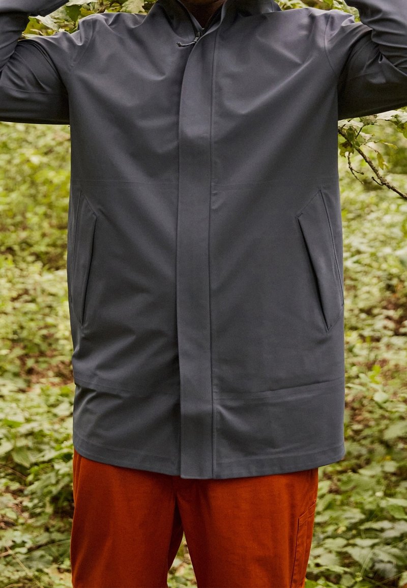 Manteau gris résistant à l'eau avec capuche, poches avant et coupe droite. Porté par-dessus un pantalon orange, avec une texture lisse et un design simple.