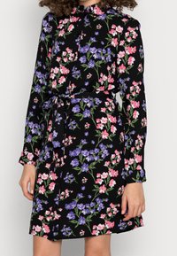 Květinové šaty s dlouhými rukávy z černé látky, zdobené růžovými a fialovými květinami, s límečkem, zapínáním na knoflíky a odpovídajícím opaskem v pase.