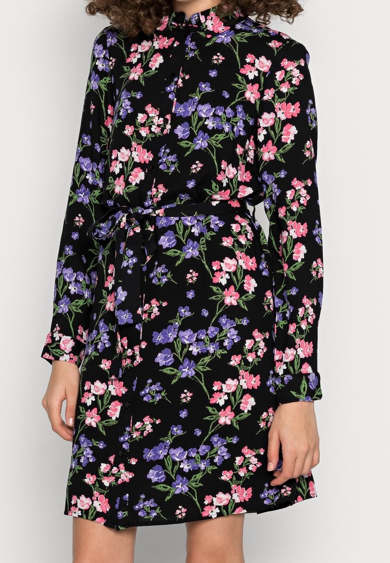 Květinové šaty s dlouhými rukávy z černé látky, zdobené růžovými a fialovými květinami, s límečkem, zapínáním na knoflíky a odpovídajícím opaskem v pase.