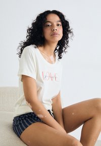 Camiseta de manga curta de cor creme clara com o texto "AMA-te a ti próprio" em coral e azul. Combinada com calções às riscas azul-marinho, apresentando um corte descontraído.