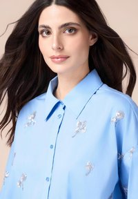 Helles blaues Hemd mit Button-Down-Kragen, verziert mit silbernen Paillettenblumen-Stickereien auf dem Stoff. Glatte Textur und entspannte Passform.