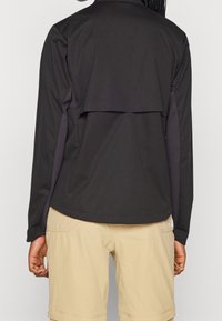Giacca nera a maniche lunghe con design posteriore a strati, caratterizzata da una texture liscia e pannelli sulle spalle in contrasto, abbinata a pantaloni corti beige chiaro.