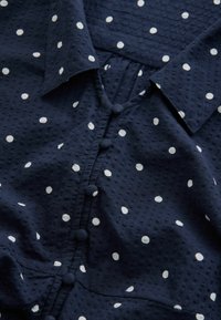 Bluse aus marineblauem Stoff mit weißen Polka-Dots, mit einem Kragen, strukturierter Oberfläche und Knopfverschluss entlang der Vorderseite und Passe.