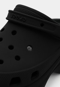 Tamanco preto de plástico com orifícios de ventilação, sola texturizada e parte superior lisa. Apresenta um rebordo elevado e o logótipo da marca na tira.