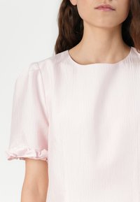 Blusa rosa clara em tecido enrugado com decote redondo e mangas curtas, apresentando detalhes de franjas nas bainhas. Textura suave.