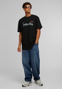 Sort T-shirt med blå og rød grafisk tekst "Members Only" og "WORLDWIDE." Parret med løse blå denimjeans og sneakers i blandede farver.
