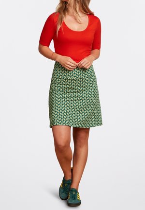 Femme portant un haut rouge à manches courtes, une jupe verte à motif et des baskets vertes avec des rayures jaunes, debout, les mains jointes.