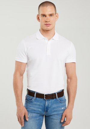 Gespierde man met kort haar, gekleed in een wit poloshirt, blauwe spijkerbroek en een donkerbruine leren riem, staand tegen een effen achtergrond.