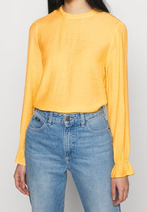 Blusa gialla a maniche lunghe con finitura texturizzata, collo rotondo e polsini arricciati, abbinata a jeans in denim blu chiaro.